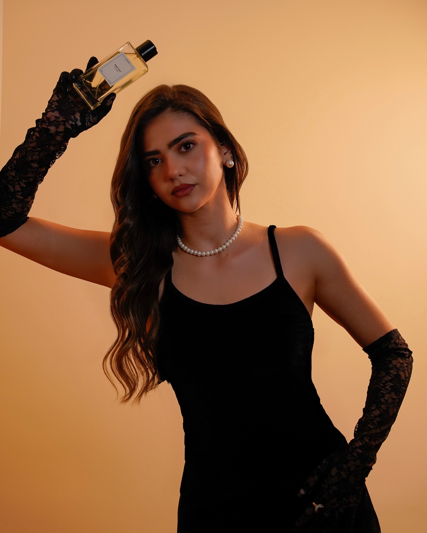 Woman in a black dress and gloves posing with a perfume bottle against a warm studio background for luxury fragrance photography.